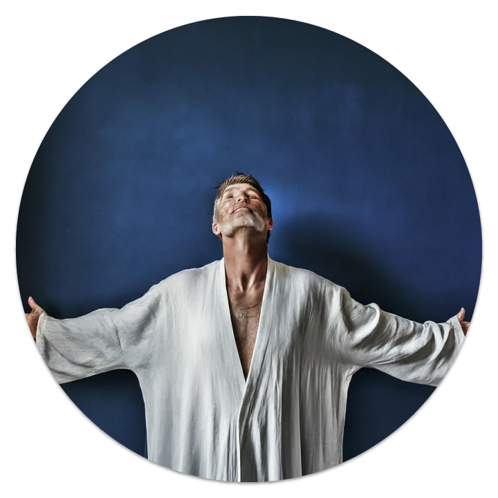 A good looking man, mid-40s, against a deep blue, background with stars, dressed in white robes, his arms outstretched, head tilted upwards