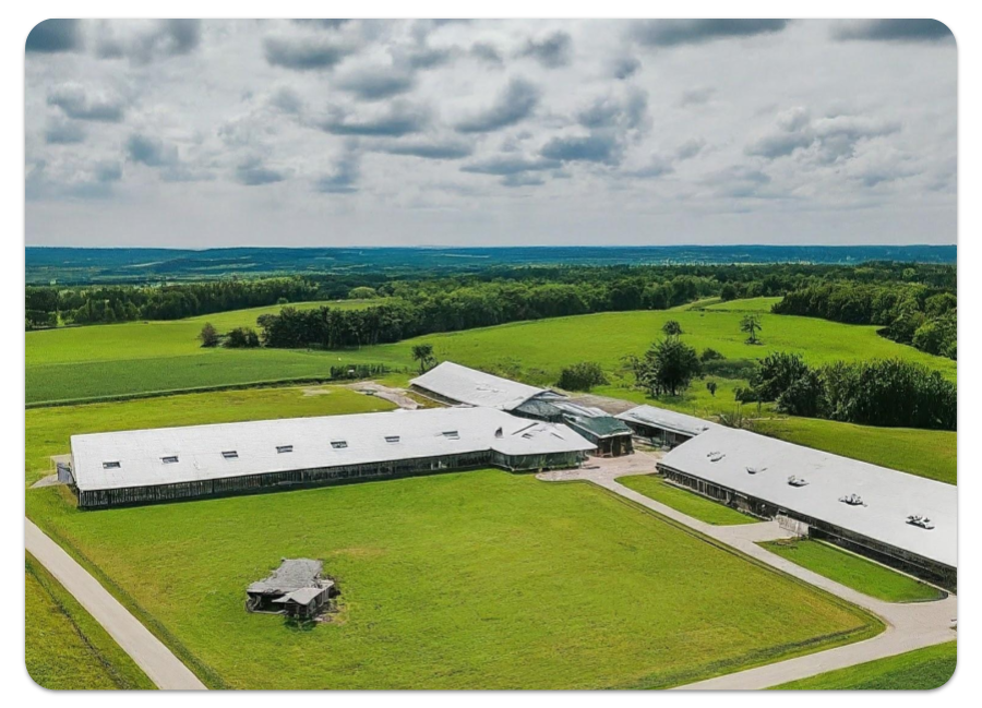 A picture of a long a sprawling farm taken from a drone. The buildings are more like dormitories rather than barns, and there is a fancy main office building. We can see various fields and some forested areas too.